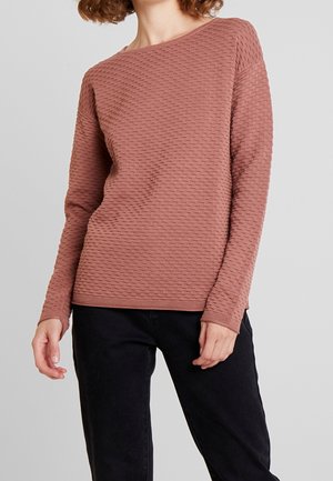 Frau trägt ein strukturiertes, staubig rosa langärmeliges Oberteil mit Bootsausschnitt und schwarze Hose vor einfachem hellem Hintergrund.