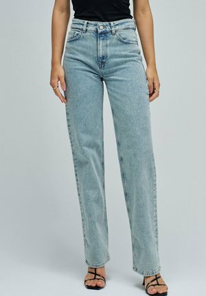 Femme portant un jean taille haute bleu clair à jambes droites, un haut noir sans manches et des sandales à talons ouvertes et à lanières noires, debout.