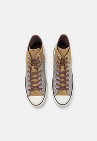 Wysokie trampki z brązową cholewką z płótna z teksturowanymi paskami i brązowymi sznurowadłami. Biała gumowa czubek buta i czarny pasek wykończenia.