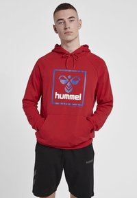 Röd hoodie med en blå fyrkantig grafisk design som har märket "hummel" i vit text. Har en dragsko till huvan och en framficka.