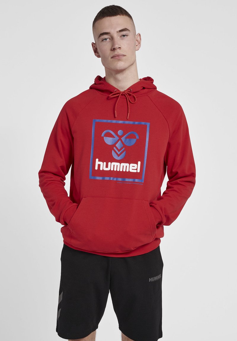 Röd hoodie med en blå fyrkantig grafisk design som har märket "hummel" i vit text. Har en dragsko till huvan och en framficka.