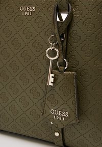 Oliven grøn håndtaske med præget diamantmønster, der har guld "GUESS 1981" logo, nøglecharme og sorte detaljer. Struktureret overflade og hardware-detaljer.