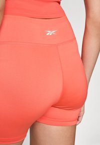 Coral fabric shorts with a high waistband, featuring a white logo. Smooth texture and close-fitting design.