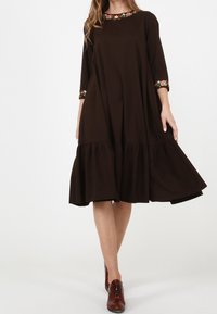 Kvinna i en lös mörkbrun klänning med blommönster på kragen och ärmarna, kombinerad med bruna läder snörskor.