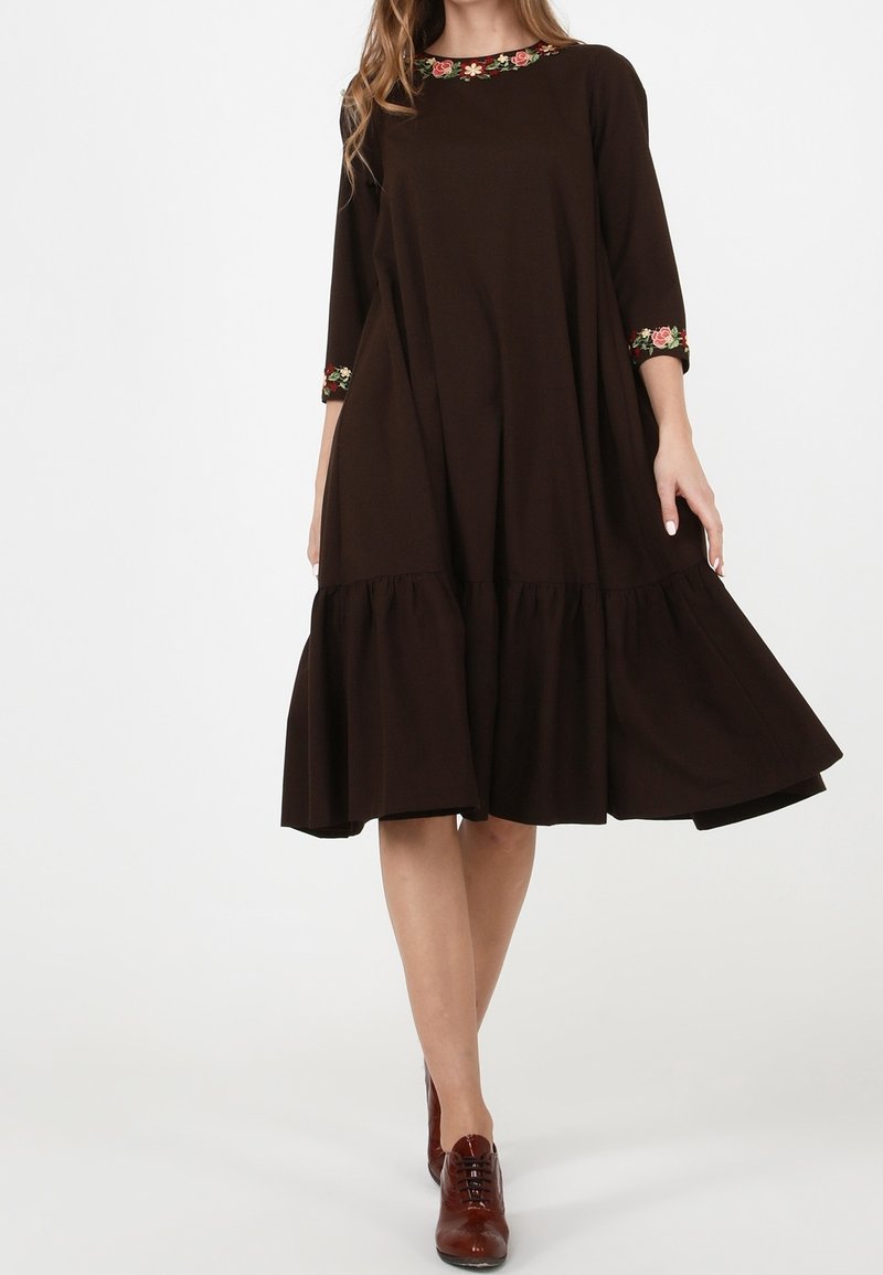 Kvinna i en lös mörkbrun klänning med blommönster på kragen och ärmarna, kombinerad med bruna läder snörskor.