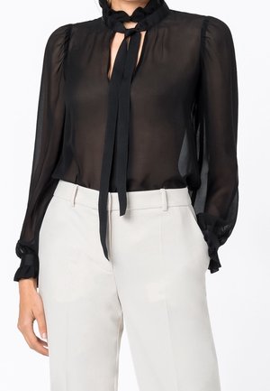 Femme portant une blouse noire transparente à manches longues avec un nœud au cou et des poignets à volants, associée à un pantalon blanc taille haute.