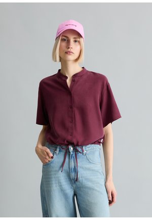 Kobieta ubrana w bordową bluzkę na guziki z krótkim rękawem i ściągaczem w talii, jasnoniebieskie dżinsy oraz różową czapkę z czarnym napisem.