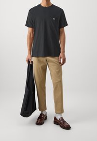 Dark gray cotton t-shirt with pocket, tan chinos, brown loafers, and black outer jacket held in hand. Simple design, no patterns or accents.