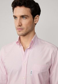 Camisa de cuadros de gingham en rosa claro con cuello abotonado, un bolsillo en el pecho y botones blancos. Textura de tela suave y corte entallado.