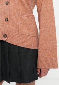 Rustinvärgad cardigan med en texturerad stickning, med knappfront och en enkel ficka. Matchas med en veckad svart kjol.
