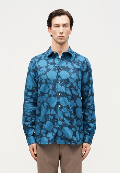 Homme portant une chemise à manches longues bleue à boutons avec un grand motif floral et un pantalon marron, debout devant un fond blanc.
