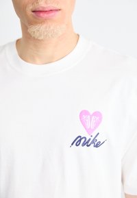 Camiseta de algodón blanca con un gráfico de corazón rosa con un diseño de zapatilla y texto "mike" en azul marino. Cuello redondo sencillo y corte relajado.