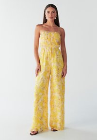 Geltonas be petnešėlių kombinezonas su sugriebtu liemeniu ir plačiomis kelnėmis. Visame audinyje gėlių raštas; lengvas ir laisvai krentantis audinys.