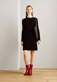 Juoda šventinta megzta suknelė su ilgomis rankovėmis, iki kelių ilgio, derinama su blizgiais raudonais kulkšnių batais. Modelis laiko juodą rankinę.