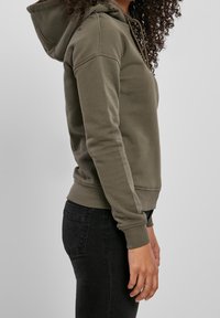 Sweat à capuche vert olive avec poche avant, poignets et ourlet côtelés, coupe décontractée, porté avec un jean foncé.