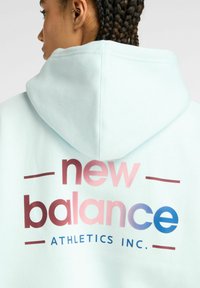 Sudadera azul claro con un gran logotipo en degradado que dice "new balance" en rosa y azul, y "ATHLETICS INC." en negro. Acabado de tela texturizada.