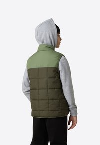 Vadderad väst med en grön överdel och en mörk olivgrön nederdel. Har en ståkrage och rektangulär quiltad textur. Bärs över en grå hoodie.