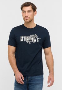 Navyblaues Baumwoll-T-Shirt mit einer silbernen Grafik eines laufenden Pferdes und dem Wort "MUSTANG" in weißer Schrift. Kurze Ärmel und Rundhalsausschnitt.