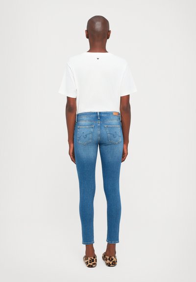 Tunna blå denimjeans med subtil blekning, i kombination med en enkel vit t-shirt och leopardmönstrade ballerinas, sedd bakifrån.