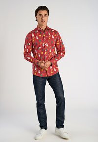 Rode button-up shirt met een patroon van sneeuwpoppen, ornamenten en bladeren, gecombineerd met donkere jeans en witte sneakers.