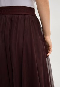 Woman wearing a dark burgundy mesh skirt with an elastic waistband embroidered with "JOOP!", standing against a plain background.