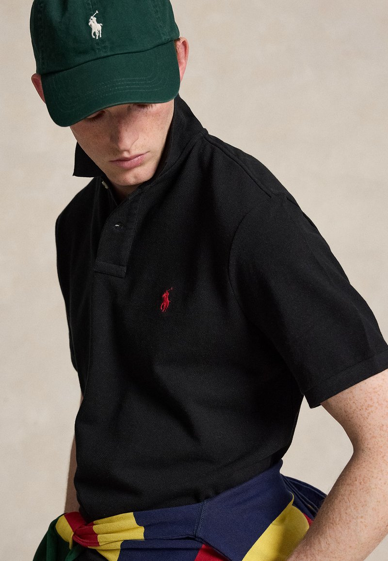 Ung mand iført en grøn kasket, sort poloshirt med rødt logo og farverig jakke bundet om taljen, kigger nedad.