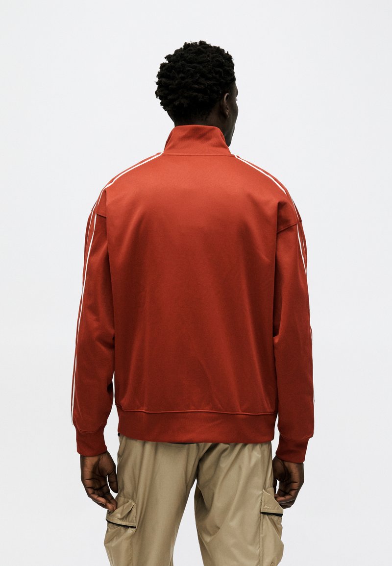 Uomo con capelli ricci scuri che indossa una giacca sportiva rossa con strisce bianche e pantaloni cargo beige, di spalle su uno sfondo bianco uniforme.