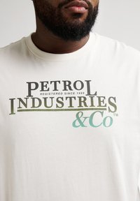 Camiseta blanca de algodón con el logo "PETROL INDUSTRIES & Co" en verde oscuro y azul verdoso. Diseño de cuello redondo, textura suave y corte relajado.