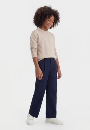 Niño sonriente con cabello rizado, vestido con un suéter beige, pantalones anchos azul marino y zapatillas blancas, de pie contra un fondo liso.