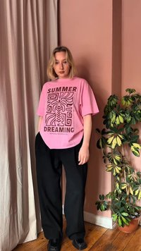 Ung kvinne som står innendørs iført en rosa T-skjorte med teksten «Summer Dreaming», svarte bukser, svarte sko, ved siden av en grønn, flerfarget potteplante.