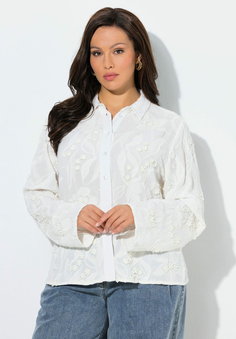 Kvinde med langt mørkt hår iført guldringe i ørerne og en hvid knapbluse med blomsterbroderi samt blå jeans.