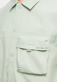 Ανοιχτό πράσινο πουκάμισο με κουμπιά, φτιαγμένο από μαλακό ύφασμα, διαθέτοντας τσέπη στο στήθος με καπάκι και το λογότυπο "OAKLEY" κεντημένο σε ανοιχτό μπλε.