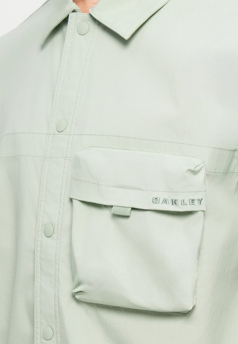 Camisa de botón en verde claro, hecha de tela suave, con un bolsillo en el pecho con tapa y "OAKLEY" bordado en azul claro.