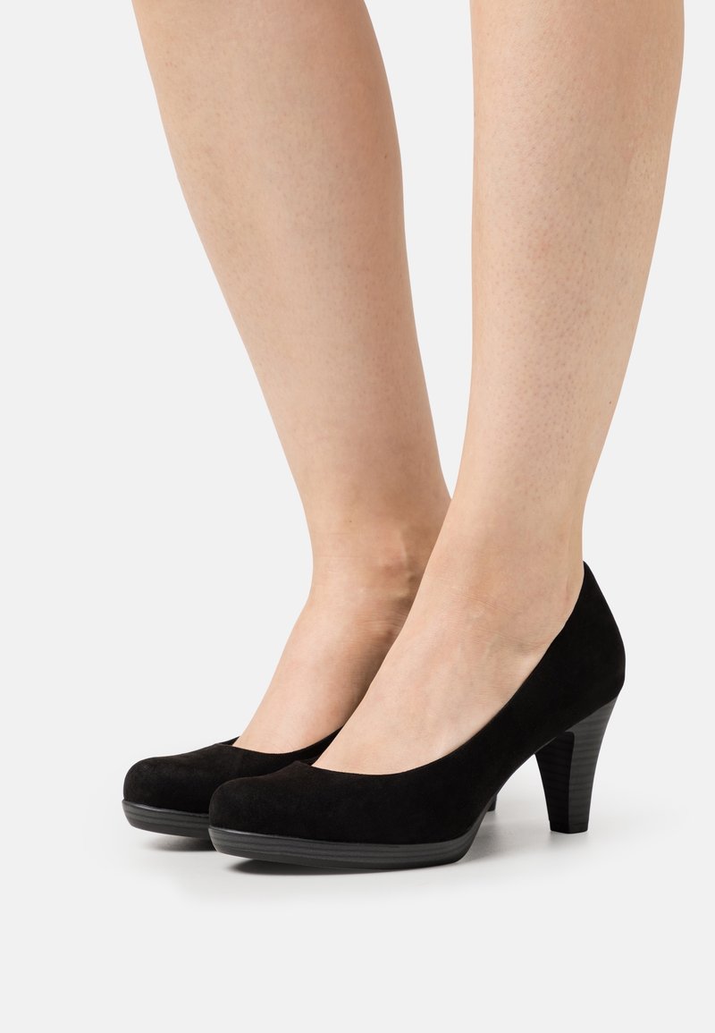 Des pieds portant des chaussures à talons moyens fermées en daim noir avec un bout rond et des talons empilés sur un fond blanc.