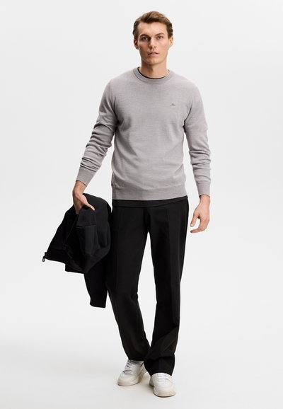 Νεαρός άνδρας όρθιος, φοράει ανοιχτό γκρι πουλόβερ, μαύρο παντελόνι, λευκά αθλητικά παπούτσια, κρατώντας ένα μαύρο μπουφάν στο αριστερό του χέρι.