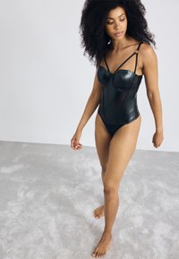 Vrouw met krullend haar die blootsvoets op een lichtgekleurde vloer met tapijt loopt, gekleed in een zwarte leren bodysuit met dunne bandjes.