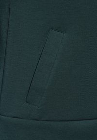 Tessuto di felpa verde scuro con una superficie liscia, dotato di una tasca laterale discreta e di una vita a costine. Design minimalista.