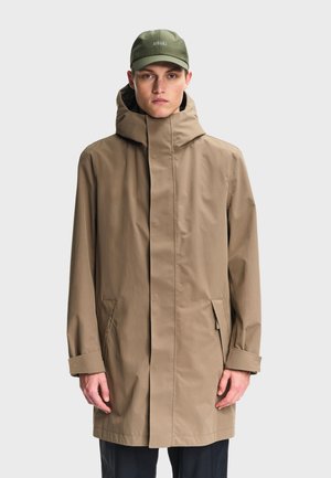 Man wearing a beige hooded raincoat with front pockets and an olive green cap, standing against a plain light background.