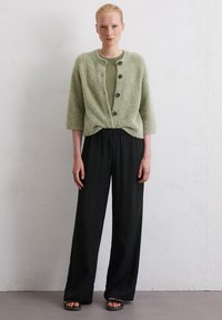 Lichtgroene, fuzzy cardigan met driekwartmouwen, voorzien van donkere knopen, gecombineerd met losse zwarte broeken en sandalen. Textuurrijke stof zichtbaar.