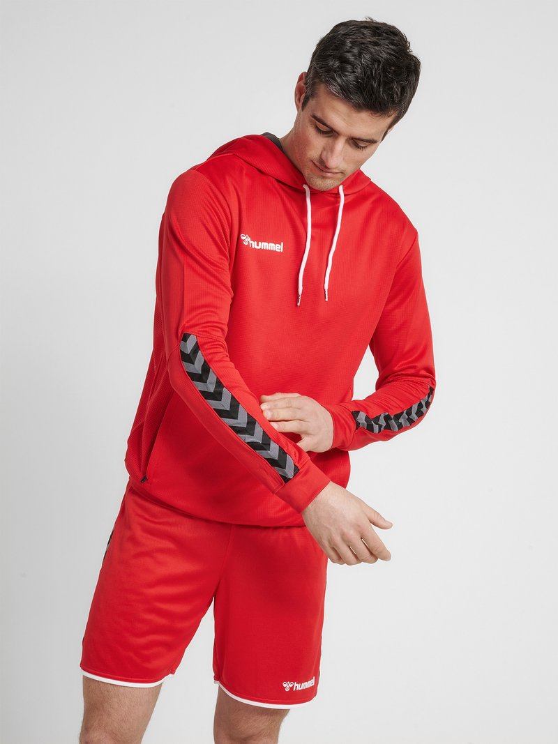 Man adjusting sleeve of red hoodie with black chevron stripes, wearing matching red shorts against plain gray background.