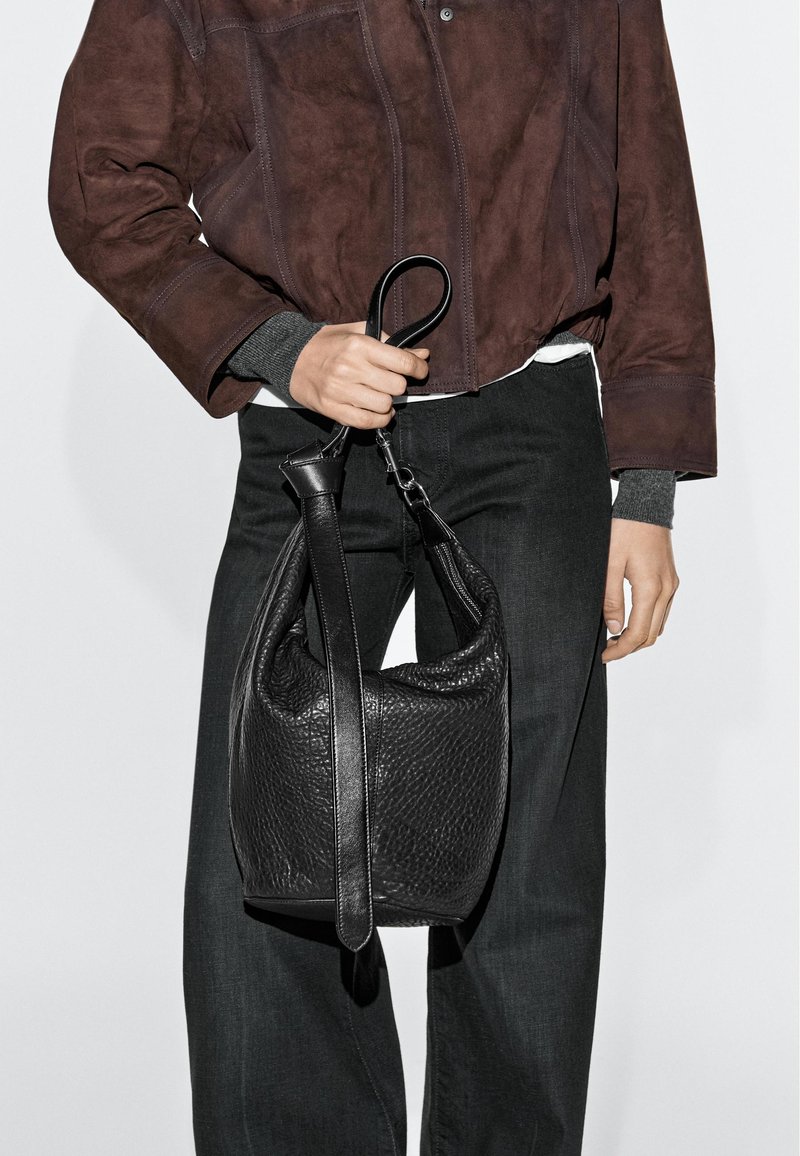 Personne portant une veste marron foncé et un pantalon noir, tenant un sac à main en cuir noir texturé par sa bandoulière.