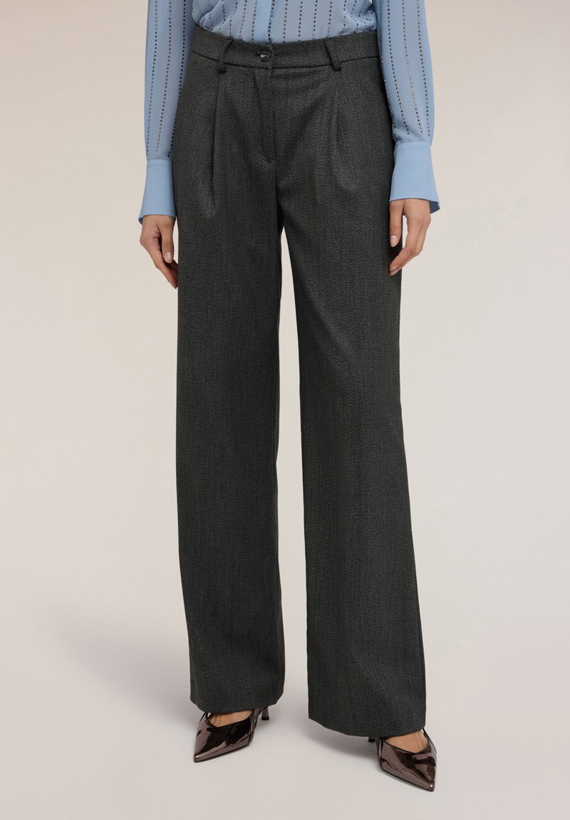 Wijdvallende broek van donkergrijze stof, met voorplooien, een knoopsluiting in de taille en een strakke uitstraling, gecombineerd met zwarte puntige schoenen.