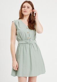 Robe vert clair avec des manches courtes légèrement bouffantes, un décolleté en V, des boutons sur le devant et une taille ceinturée, présentant une jupe fluide.