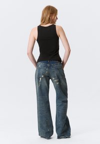 Débardeur noir associé à un jean denim bleu à jambes larges avec détails usés et éclaboussures de peinture, vu de dos.