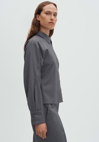 Chemise grise boutonnée avec col évasé, manches longues ornées de boutons et coupe ajustée. Texture lisse, design classique.