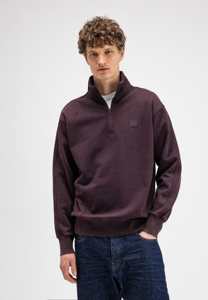 Junger Mann mit lockigem Haar, der einen dunkelvioletten Sweatshirt mit halbem Reißverschluss und blaue Jeans trägt, steht mit einer Hand in der Tasche.