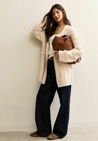 Oversized crèmekleurige gebreide cardigan met kabelpatronen, gecombineerd met een wit topje en donkere, wijde jeans. Bruine rechten suède handtas en platte schoenen.