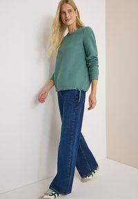 Kvinna iklädd en teal-färgad, strukturerad långärmad topp, vida blå jeans och vita sneakers med gröna ränder, gående inomhus mot neutrala väggar.