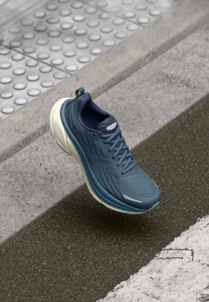 Chaussure de sport bleue avec tige en maille texturée, semelle blanche et accents verts. Caractéristiques d'un design léger et d'un matériau respirant.