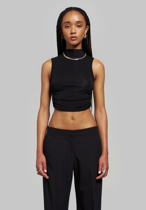 Jeune personne aux longs cheveux tressés portant un crop top noir sans manches, un pantalon noir, des boucles d’oreilles en argent en forme d’anneaux, un collier et des bagues, debout devant un fond gris.